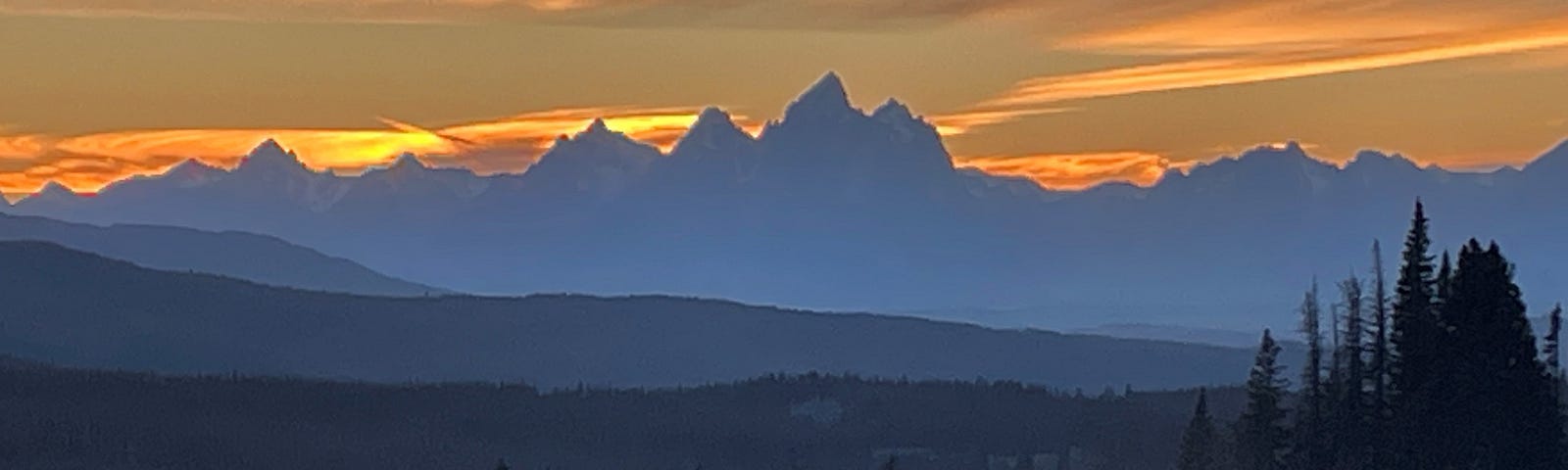 Orange and yellow sky glows brightly behind the dark silhouette of the Grand Teton mountain range.