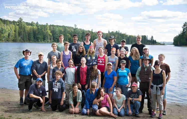 A large group of teens and a few adults gathered in front of a lake in the summer ready to go canoeing.