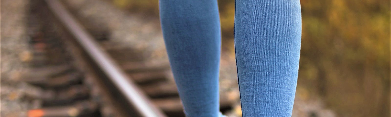 person wearing blue jeans and white tennis shoes walking on edge of railroad track