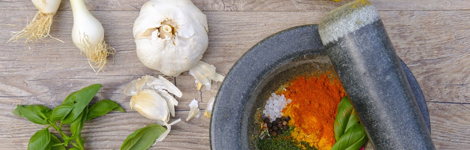 Mortar with salt, garlic, spices, and herbs, and with onion, garlic, lemons, plum cherries and basil on the table