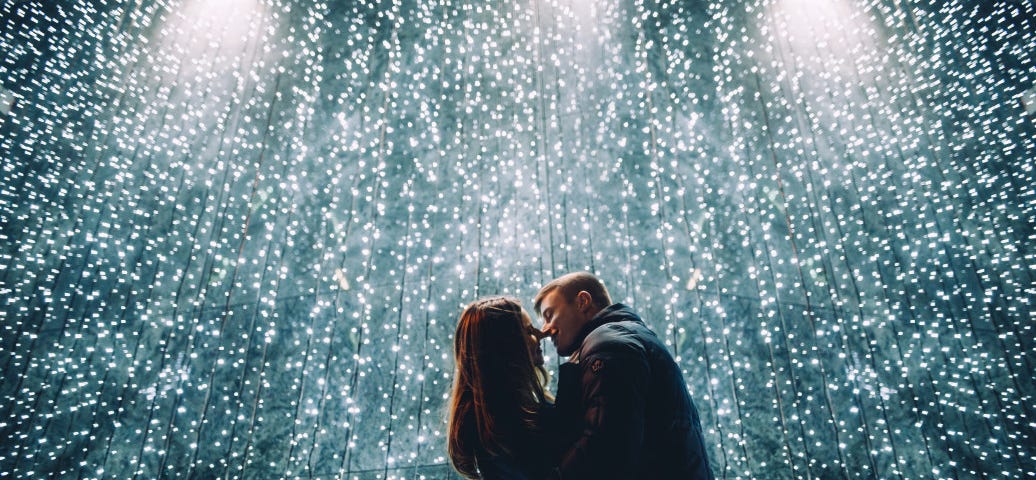 A happy couple embraces and kisses under a splay of small lights. What the 5 Love Languages Really Mean.