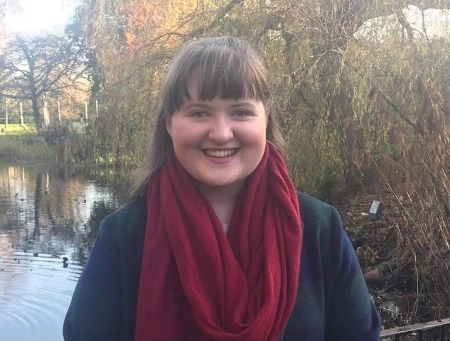 The author, smiling, wearing a red scarf, against the backdrop of a pond or stream and a thicket of trees.