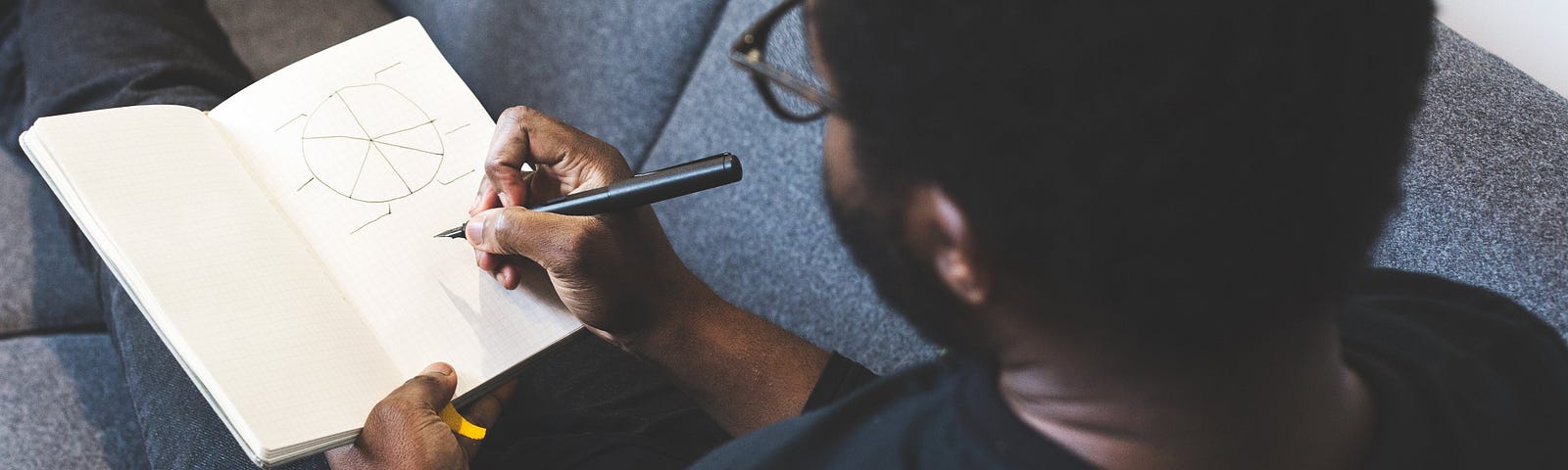 A man relaxes on the couch while he is sketching a circle in his notebook.