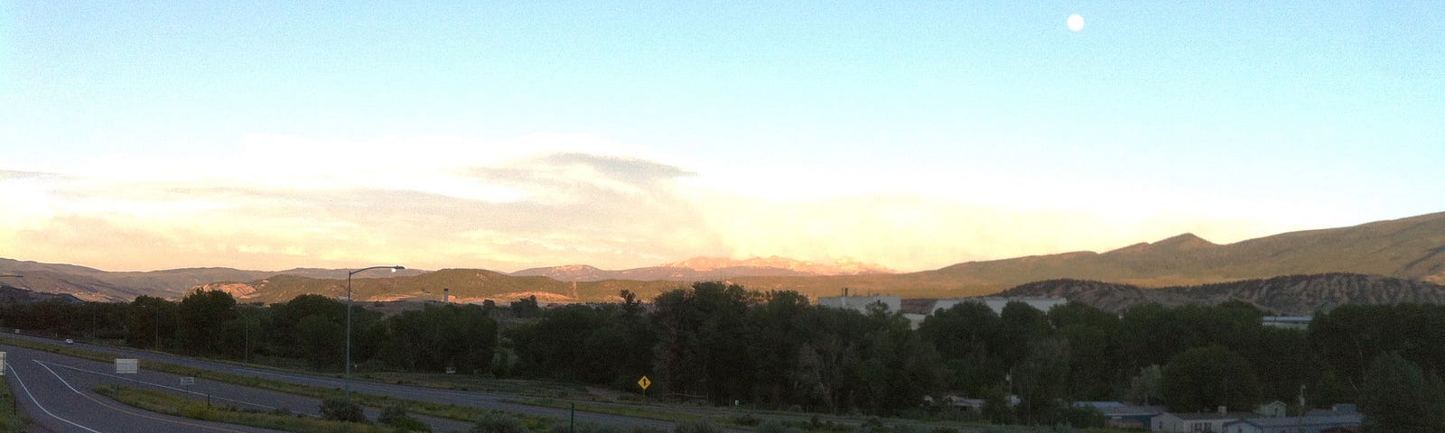 The sun goes down over the highway as the moon rises and a woman with a feathered hat and camping pack sits by the side staring off into the distance, hoping for a ride.