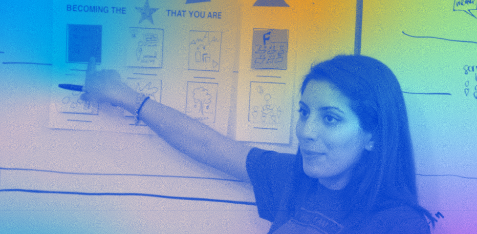 Tania Anaissie stands near a white board and points to a handout taped to its surface. The title of the handout says, “Becoming the Star That You Are.” This image is colorized with shades of light blue, gold, green and pink.