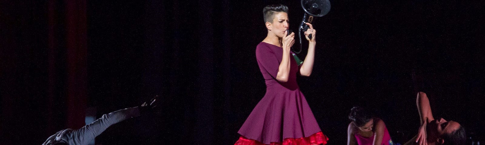 Woman in dress walking through group of performers holding a megaphone