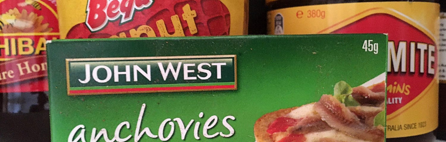 A close-up of a green rectangular box with the words, John West anchovies fillets in olive oil, on the front. Behind the box are jars of foodstuffs with yellow and red labels.