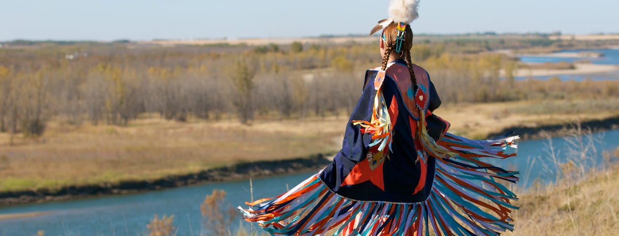 First Nations Women performing a Fancy Shawl Dance in a grass field with a river background