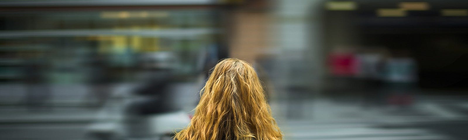 A woman with long, wavy brown hair and a dark top has her back to the camera, and is looking out onto a busy, blurred street.