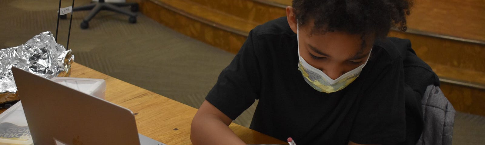 A young student sits at a computer and writes on a piece of paper. They are participating in Spark Central’s Level Up program.