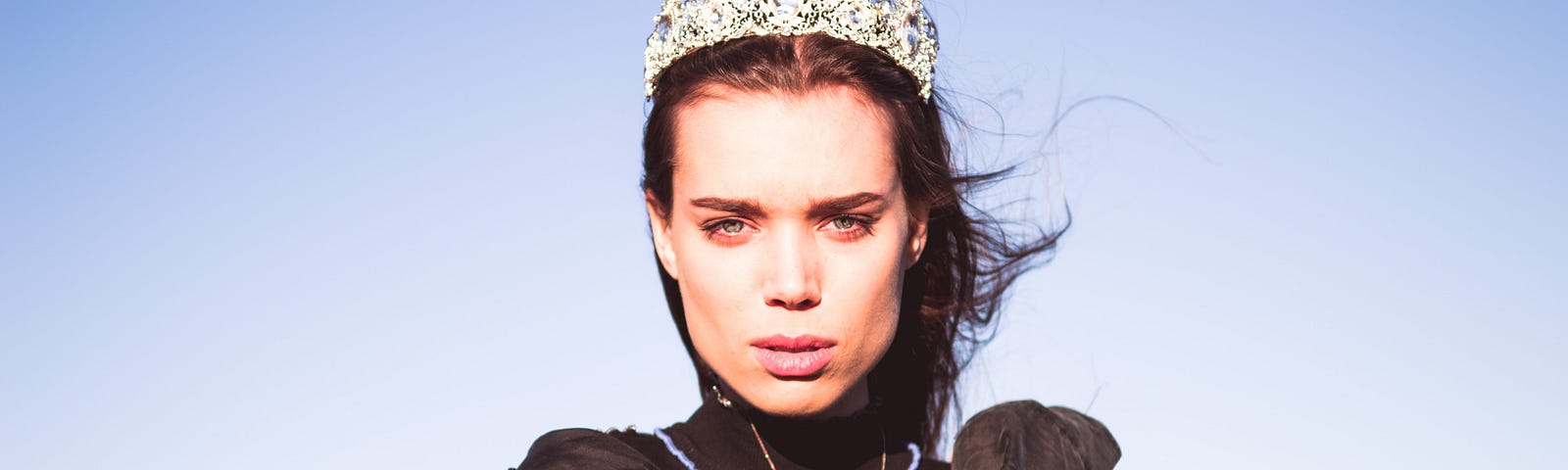 A woman wearing a black dress, a silver crown, and a gold necklace stands outside, the wind blowing her long dark hair to one side. The sky is blue behind her.