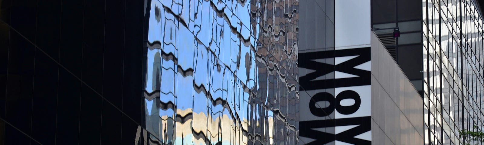 Photo of the shiny, reflecting steel front of the building of the Museum of Modern Art in New York. The sign with the vertical letters “MOMA” sticks out from the facade.