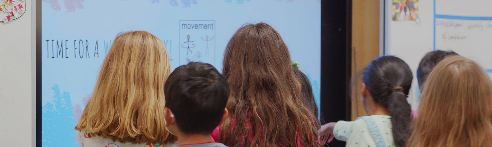 A group of elementary school children, with their backs facing the viewer, work on a large screen in their classroom.