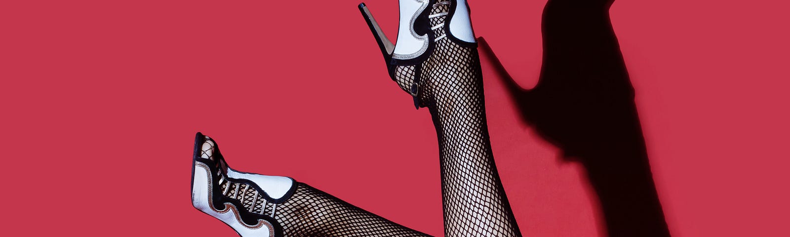 Women’s legs in fishnet stockings and black and white stilettos in front of a red background.