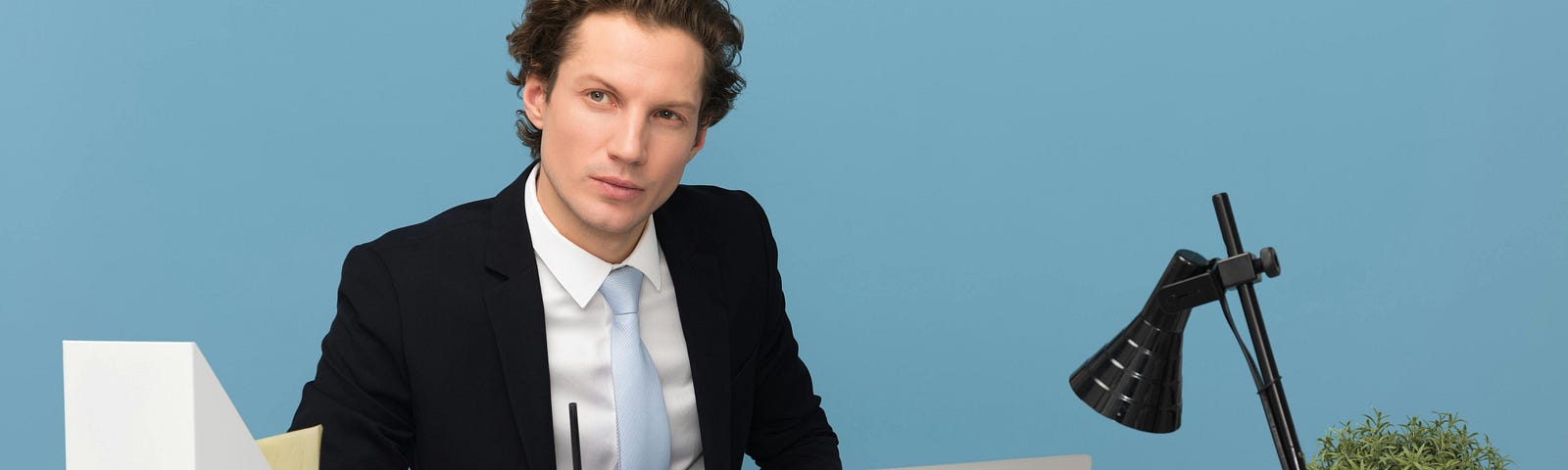 Dark-haired man dressed in black suit with white shirt and blue tie, sitting at a modernist desk with laptop, black lamp, and plant, against a bright blue background.