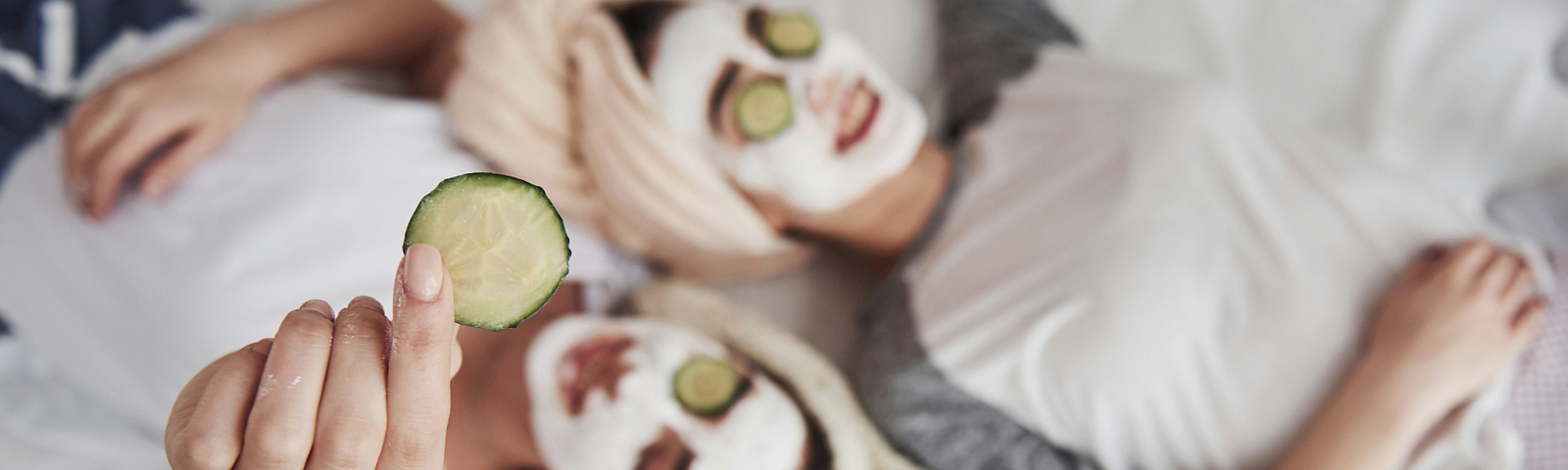 Two women lie on a bed with hair in a towel and white face-mask on their faces. They have cucumbers on their eyes and one woman holds up a slice.