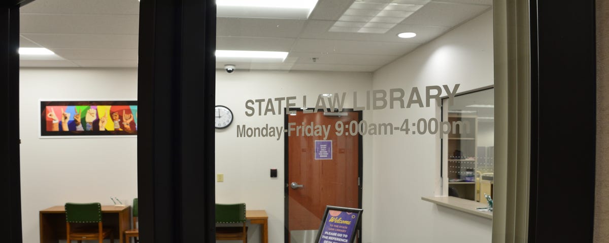 The library research room is shown from the Town Center 3 lobby. Desks and chairs are visible, as well as the service window. The text on the window of the research room reads, “State Law Library, Monday-Friday, 9:00 am-4:00 pm.”