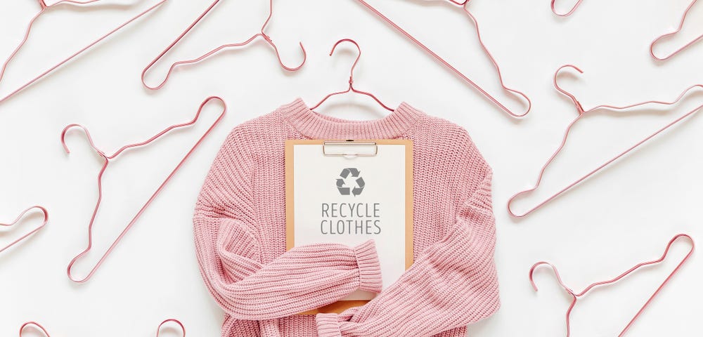 Picture of a pink pullover holding a sign with “recycle clothes” and surrounded by coat hangers.