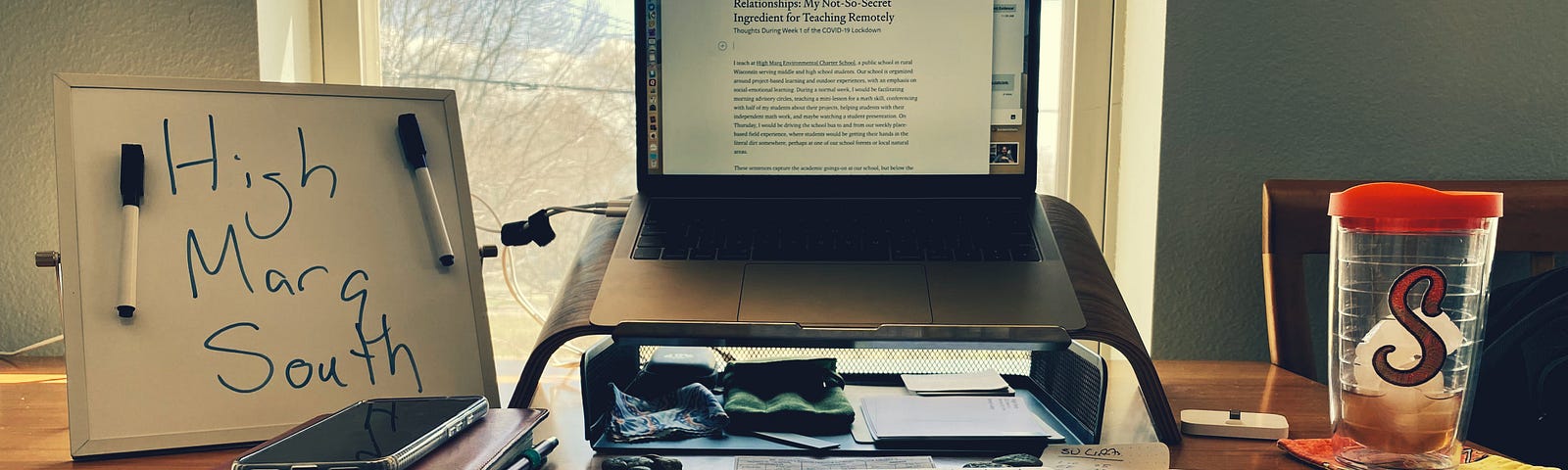 A laptop computer and various desk accessories laid out on a dining room table in front of a window