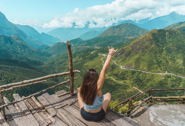 Woman sitting, back facing camera, arm raised in victory. She has climbed up to ahigh point overlooking a green mountain range. The sky is blue with white fluffy clouds.