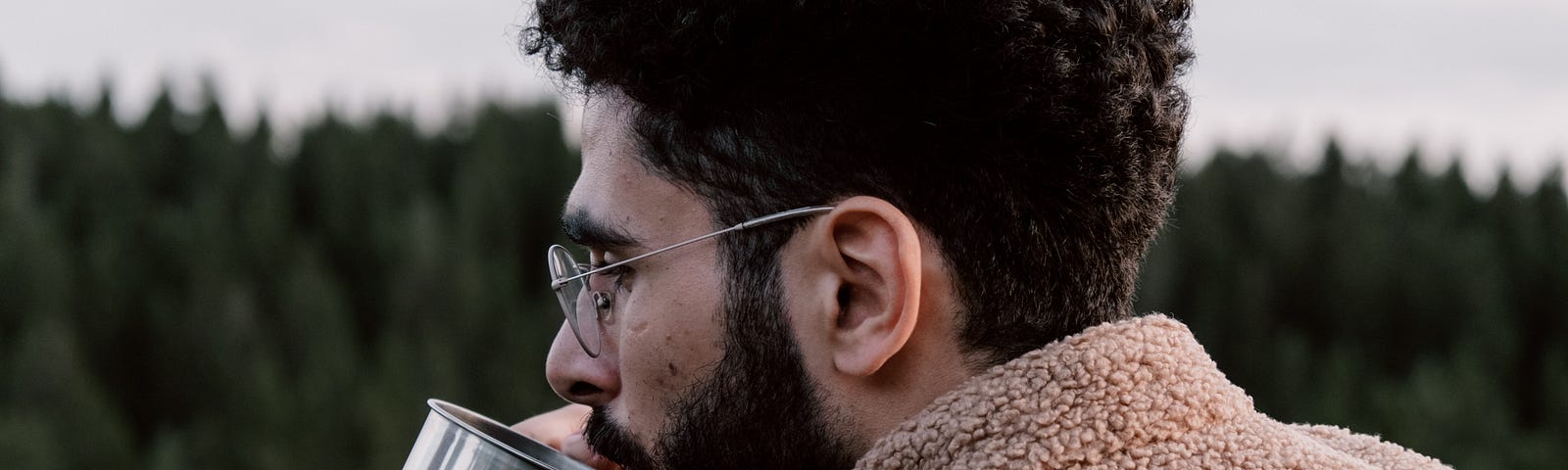 Bearded and spectacled young man sipping coffee in the backdrop of a forest