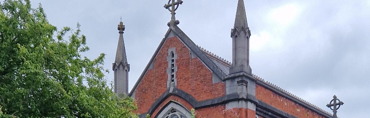A coloured image featuring the front of a red brick church with a turret on either side of a cross. The church sits behind a high stone wall and is framed by a cloudy sky.