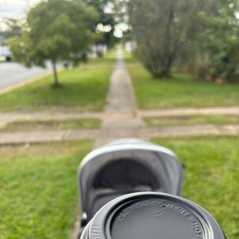A pram being pushed down the sidewalk with a coffee cup being held in a hand