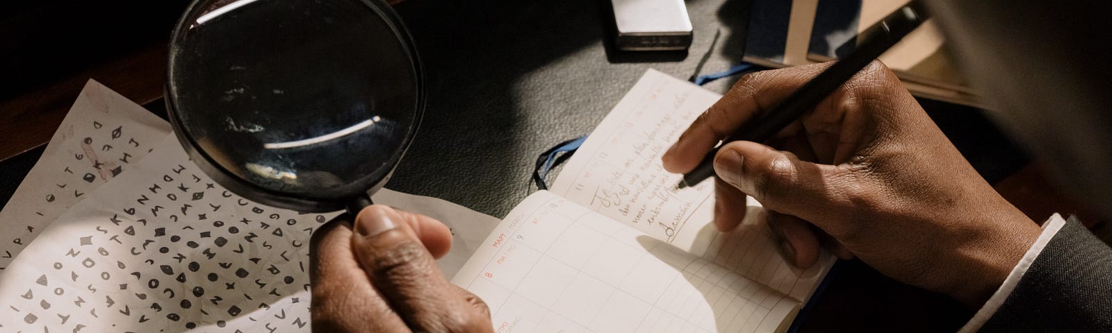 A man’s hand holding a magnifying glass in his left hand and writing down notes with his right hand in a notebook. He is looking at some coded papers.
