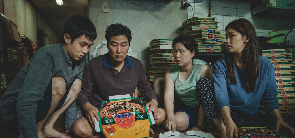 Família Kim unida com todos sentados no chão dobrando caixas de pizza. Da esq. para a dir. estão o filho, pai, mãe e filha.
