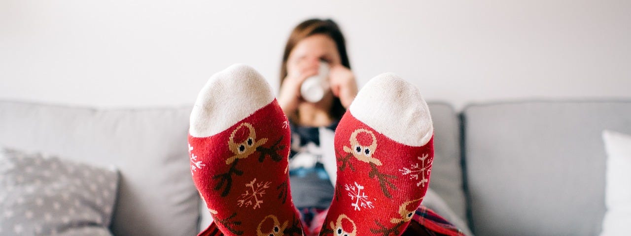 Woman sitting on a couch, drinking coffee, with sock-covered feet up on the coffee table.