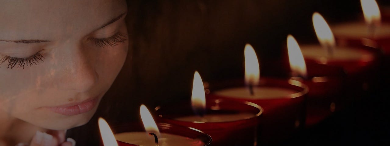 A woman’s face in the darkness, eyes closed, hands together in prayer, in front of a row of lit candles.