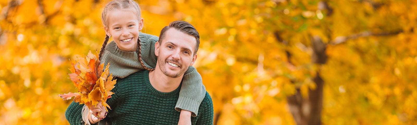 Father carries daughter through autumn trees. She holds yellow orange leaves.