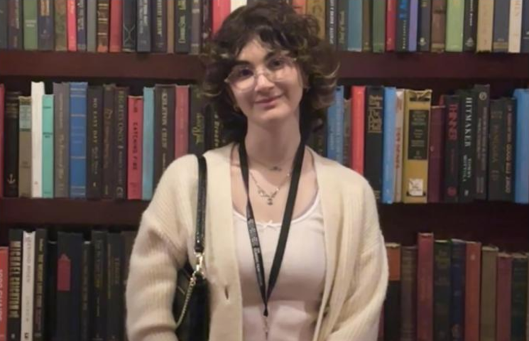 A young person wearing a pale sweater with a white shirt, glasses, black purse on their left shoulder, and a blue pass around their neck is standing in the middle of a book shelf. They have pale brown hair with some blonde streaks in the front and have a smile on.