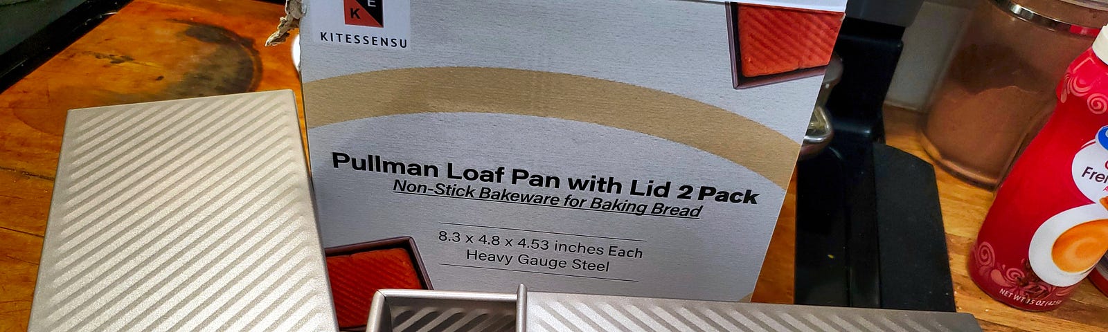 A pair of non-stick Pullman loaf pans with lids are displayed in front of their box, which claims the bakeware is ideal for baking bread. The pans appear to be new and are placed on a kitchen counter, next to a jar and a bottle.