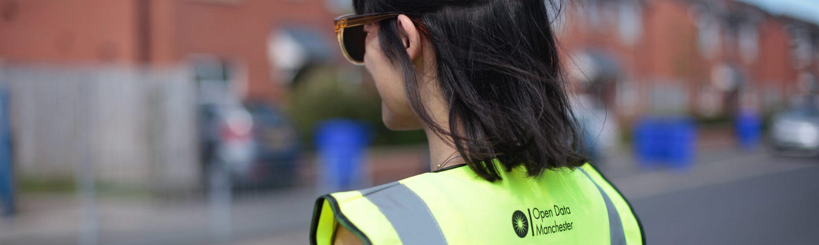 A person wearing a yellow high-visibility vest, printed with the words “Data Gatherer” and an Open Data Manchester logo, is standing on a residential street lined with red-brick houses, inputting data into a handheld electronic device.