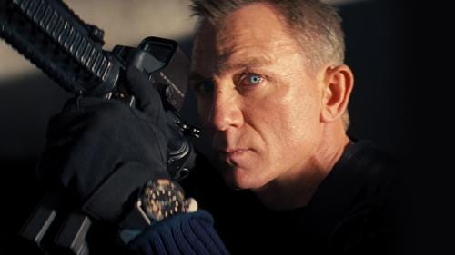 Daniel Craig a middle aged blue eyed Britain, holds up an automatic rifle at an angle with black gloves wearing a navy fisherman’s sweater. Behind him is a concrete wall in shadow. He has a look of determination on his face.