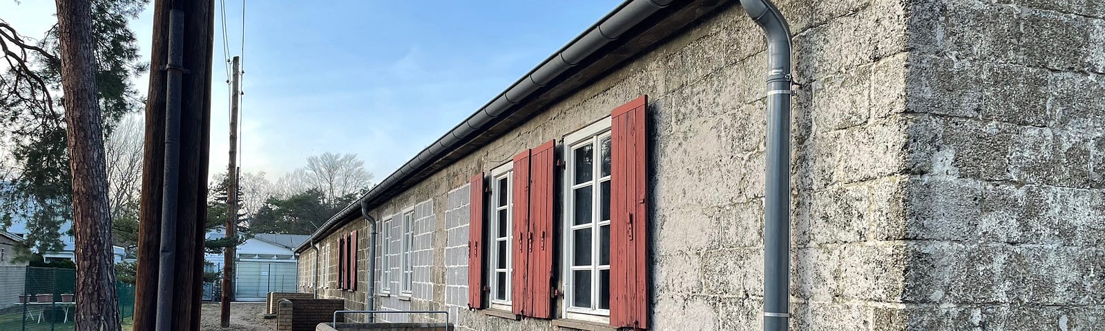 This is a photo of a cement one-story rectangular barrack with red shuttles next to its windows. The sky is blue with mature trees and telephone poles to the left.