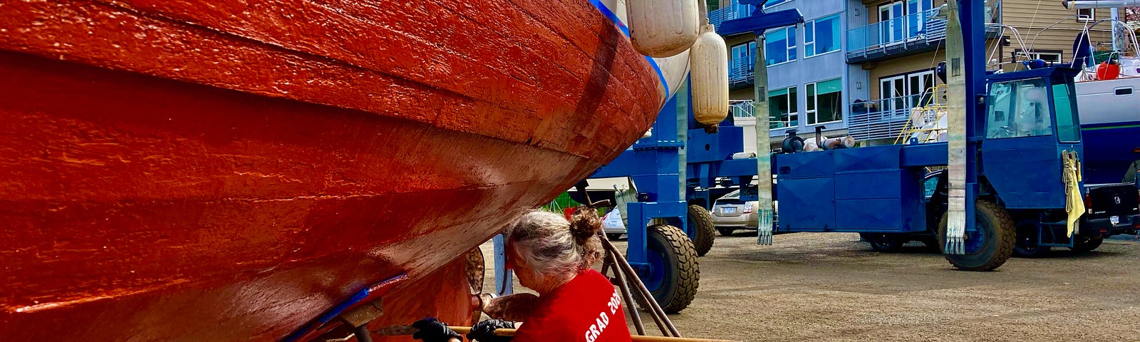 A woman is painting the hull of a boat. She’s using a roller and her hair is gray and in a bun.