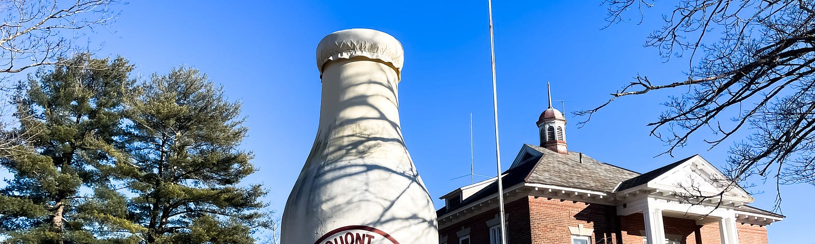 A giant stone milk bottle stands tall in the grass in front of an old brick building.