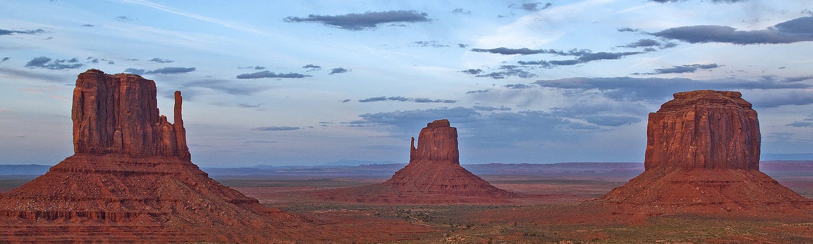Landscape photograph of three distinctive rock formations. A cluster of vast sandstone buttes underneath a blue sky with clouds.