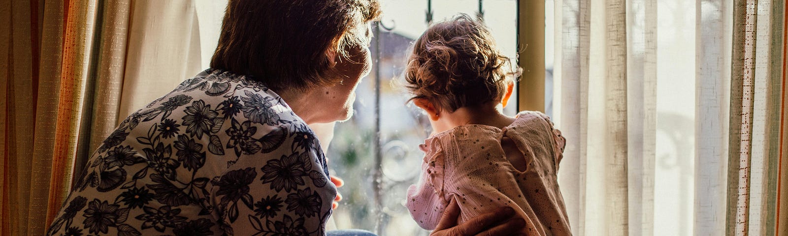 grandmother showing her granddaughter the outside world through a window and drapery…