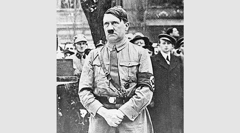 Hitler is standing in his uniform, hands folded.