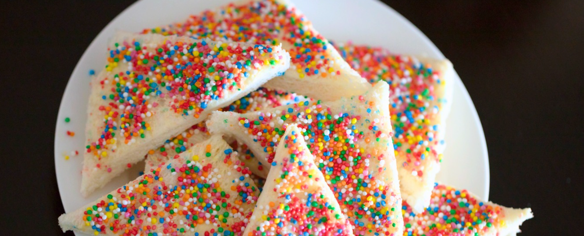 Plate of fairy bread cut into triangles. White sliced bread with 100s and 1000s rainbow sprinkles on top.