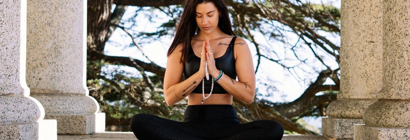 Woman sitting on a concrete outdoor space, in cross-legged, hands in prayer mode and looking slightly down. There are 4 large columns around her and a beautiful blue sky with trees in the background.