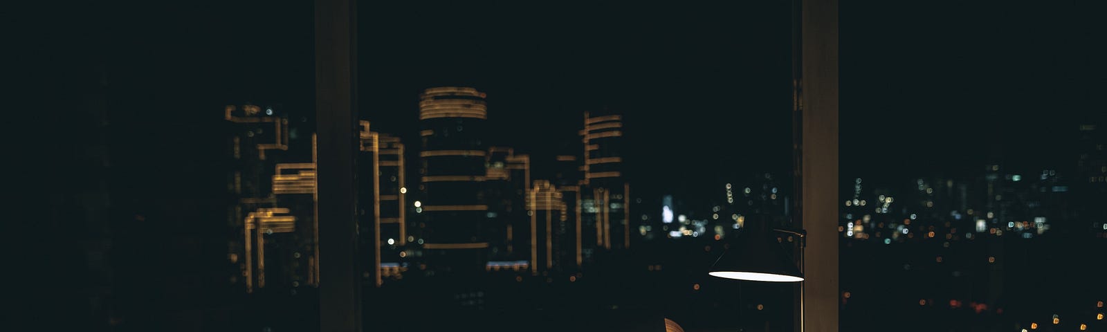 A dimly lit desk at the window of a high rise apartment.