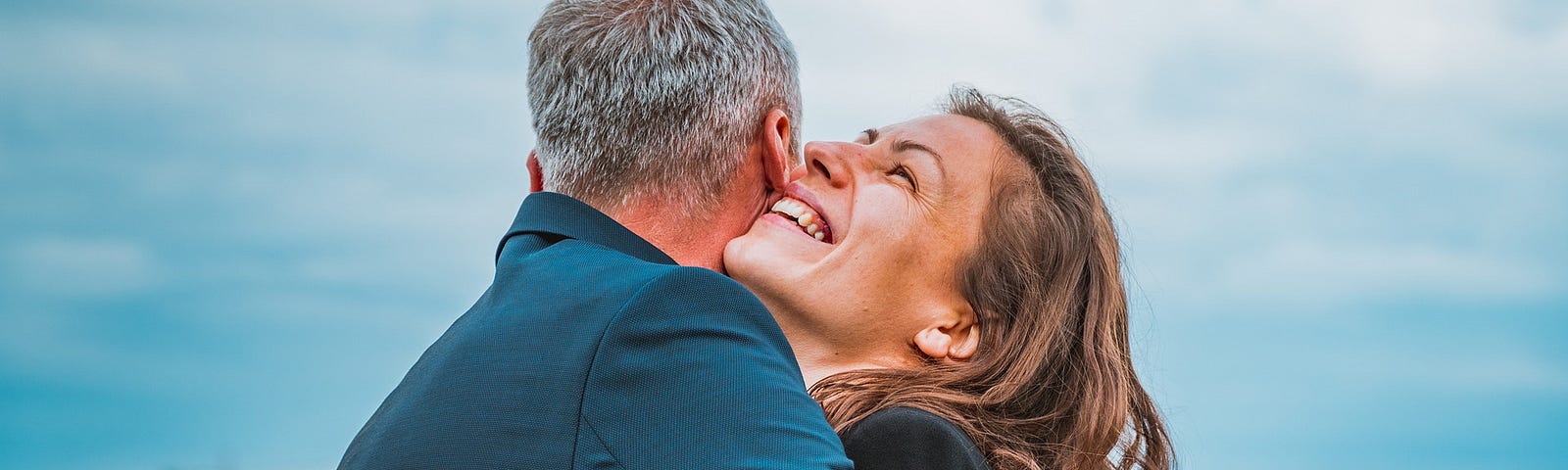 A middle-aged woman hugs a man with gray hair. She is smiling. You can’t see his face. They are outside, near water