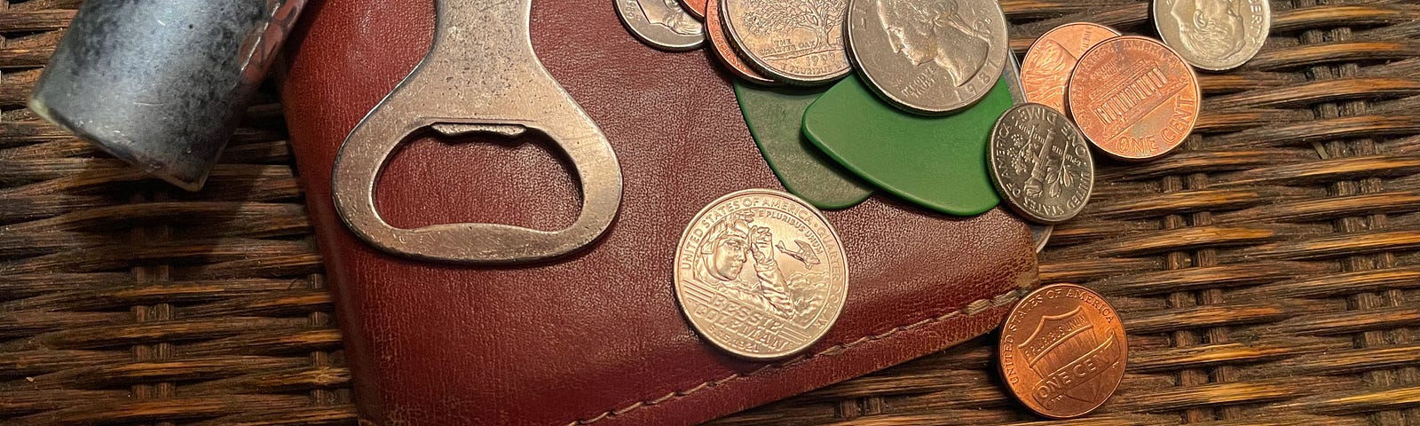 A collection of personal items on a wicker end table, including a set of keys, a tan leather pocket notebook holder, two green guitar picks, and a handful of coins, with a single quarter set aside, ready for decision-making duty.