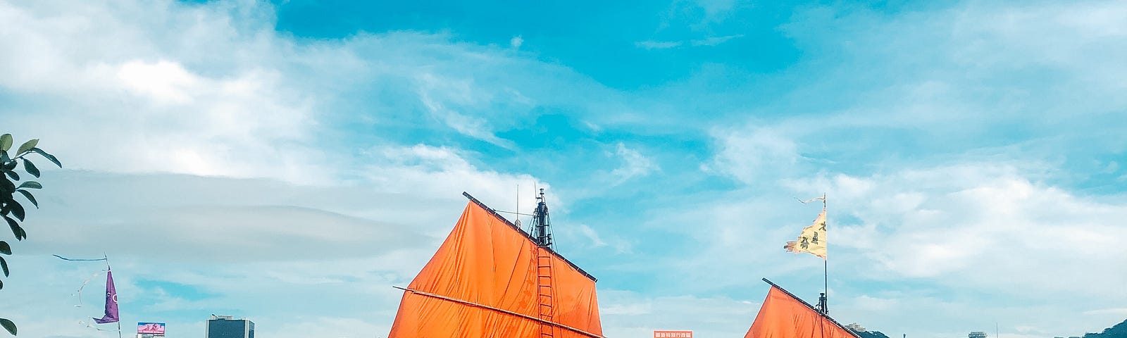 A junk rig, a traditional Chinese ship, sailing across the river connecting Hong Kong’s island from the mainland. The ship has bright orange sails that stand out from the partially cloud sky.