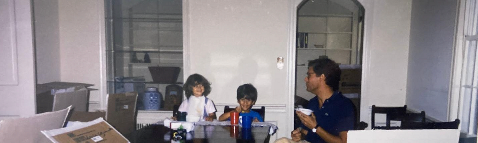 two children and one man sitting at dinner table in an empty house with moving boxes