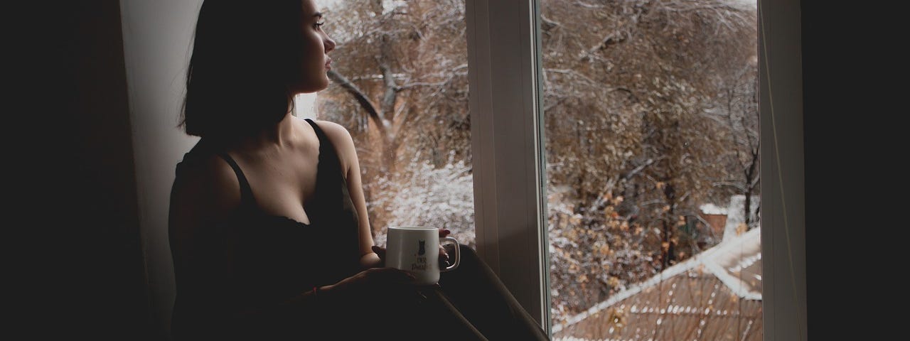 A young, white woman staring out the window in deep thought with a coffee mug in her hand.